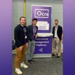 3 men standing a a conference booth next to purple banner with Ocra logo