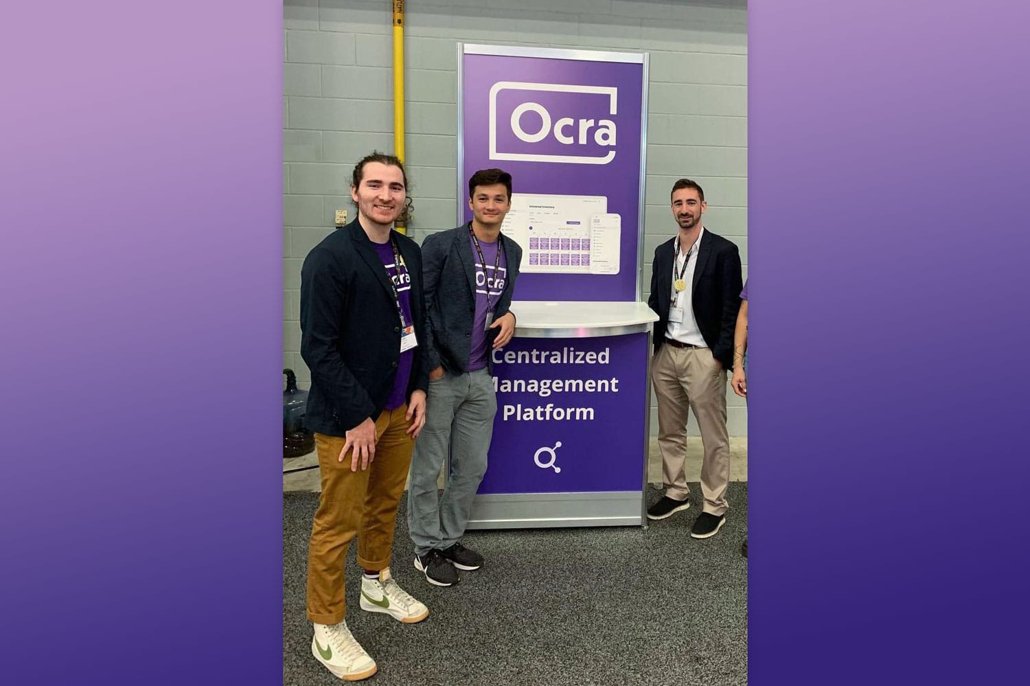 3 men standing a a conference booth next to purple banner with Ocra logo